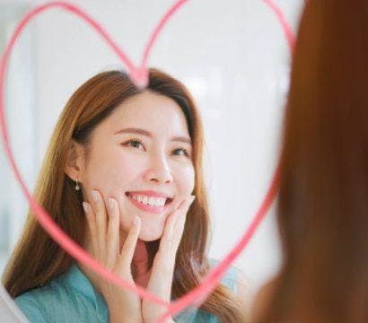 Woman in green touching her face and smiling into a mirror with a heart.