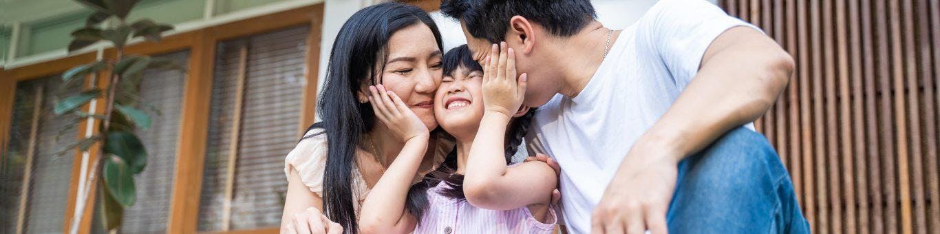 Asian mom and dad kissing their daughter while smiling.