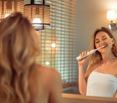 Blonde woman brushing with electric toothbrush in front of mirror.