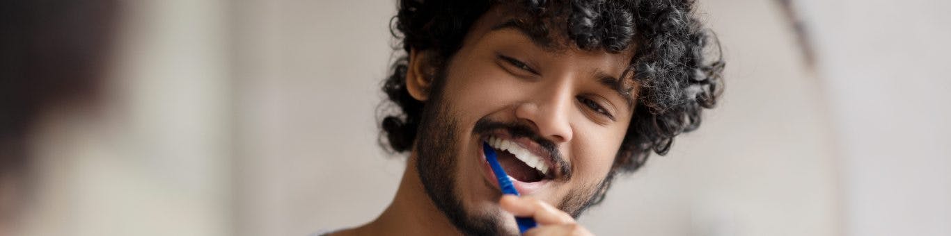 South Asian man brushing teeth in front of mirror.