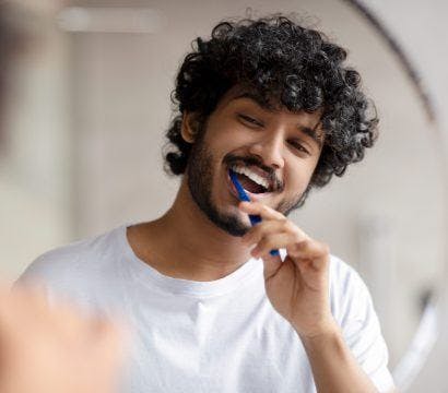 South Asian man brushing teeth in front of mirror.