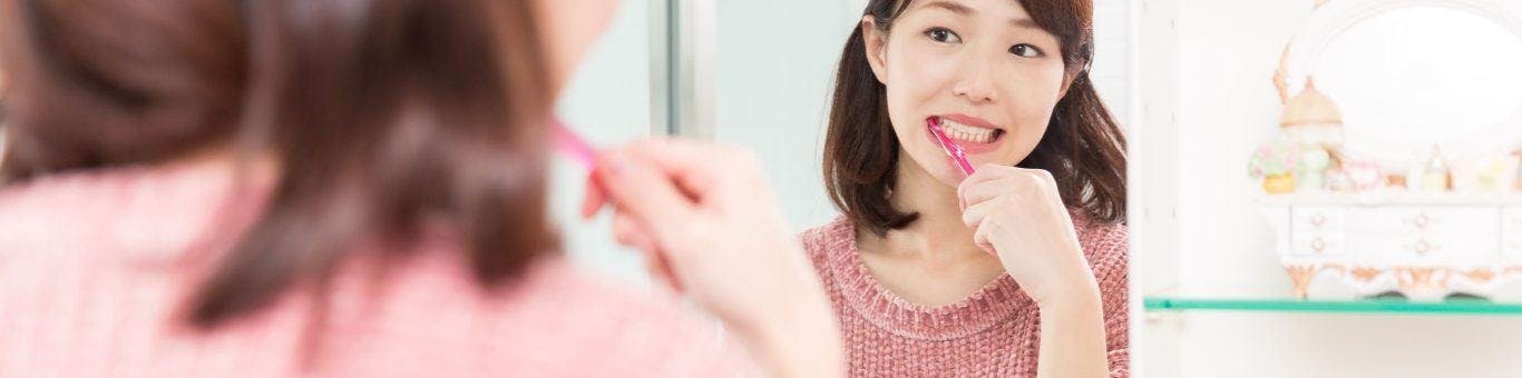 Asian woman with shoulder-length hair brushing teeth while looking at mirror.
