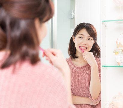 Asian woman with shoulder-length hair brushing teeth while looking at mirror.