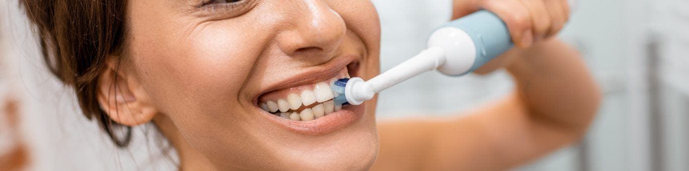 A smiling woman brushing her teeth with an electric toothbrush.
