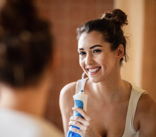 A woman holds a water floss device while smiling at the mirror.