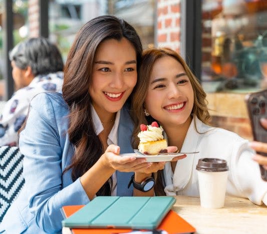 Female Asian friends smiling and taking a selfie while holding dessert outdoors.