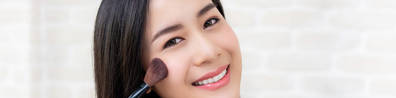 Smiling woman applying blush while holding makeup palette.