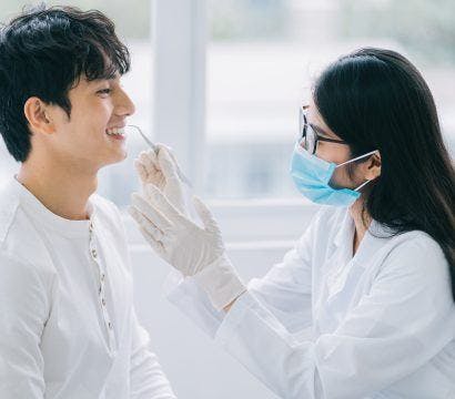 An Asian dentist checks a male patient’s teeth while he smiles.