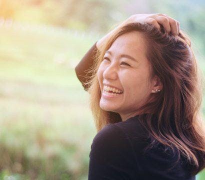 Young Asian woman runs hand through her hair and wears a big smile on her face while outdoors.