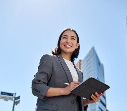 An Asian woman smiling in a business district while writing on her tablet device.