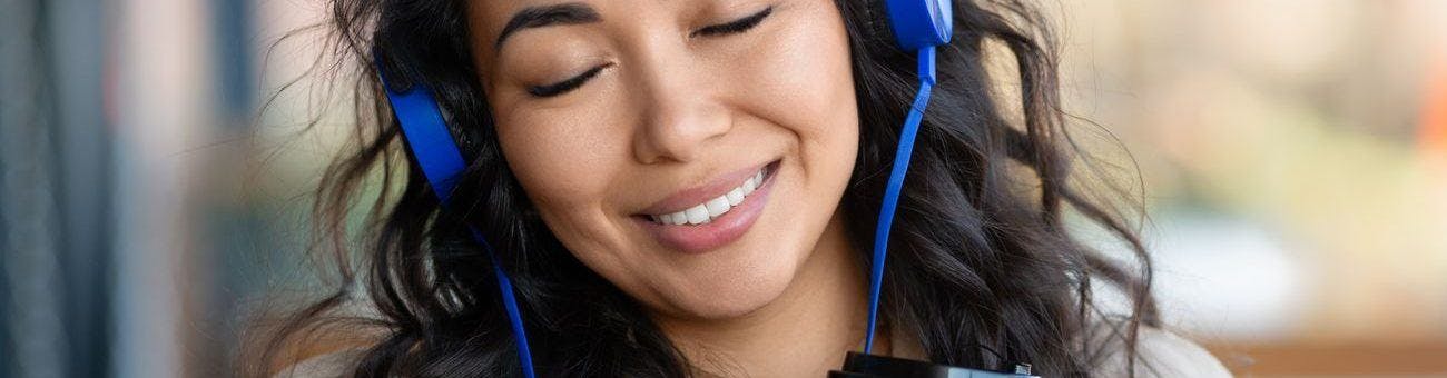 Smiling woman closing her eyes while listening to music with blue headphones and holding a cup of tea.