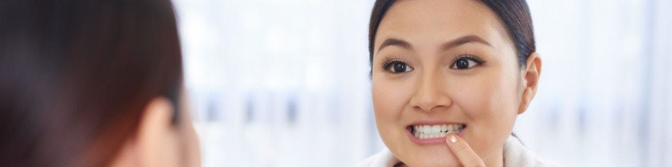 Asian woman in a white robe looking in a bathroom mirror and touching her teeth.