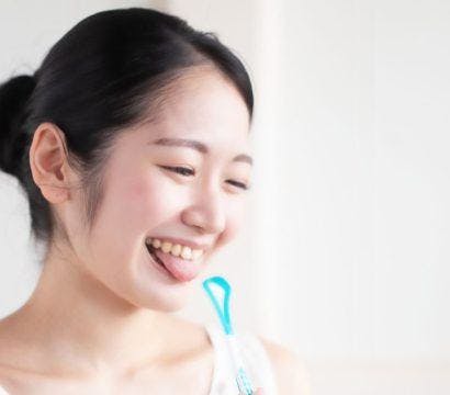 Woman in white happily holding a tongue cleaner and sticking her tongue out.