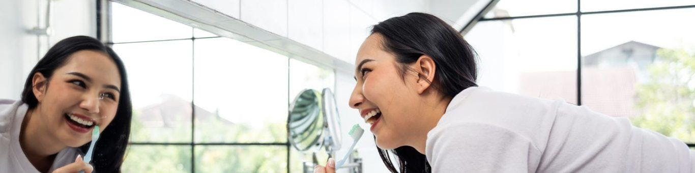 Woman brushing her teeth happily and looking in the mirror of a bright, white bathroom.