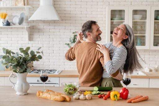 An older couple laughing and dancing in the kitchen while preparing food.