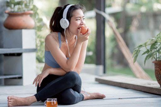 Young woman in workout clothes and headphones eating apple and drinking tea.