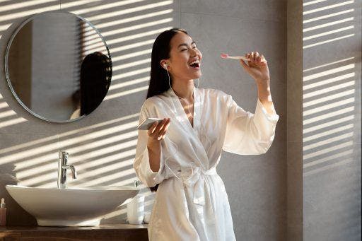 Woman in bathrobe listening to music while brushing her teeth.