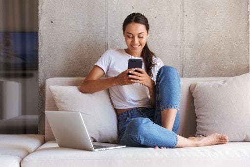 Woman on the couch with a laptop and happily looking at her mobile phone. 