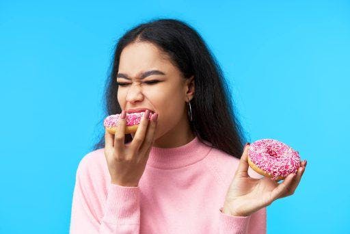 Wavy-haired mixed-race woman holding two pink doughnuts, biting into the other one.