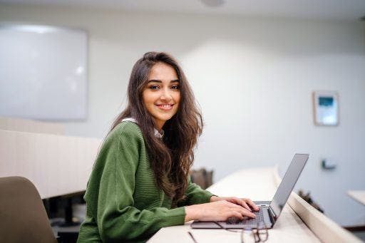 Indian or mixed Asian woman smiling while typing on her laptop in a green sweater.