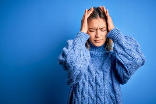 Young woman wearing a blue sweater holding her head in pain against a blue background. 