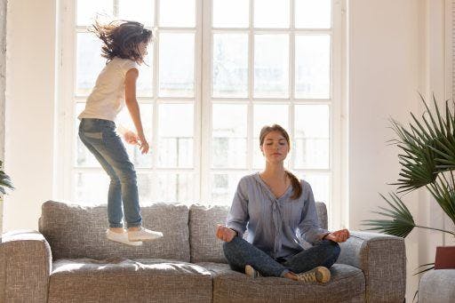 Mother doing yoga while daughter jumps on the couch. 