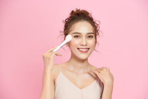Smiling woman holding a blush brush to her face with her other hand on her collarbone against a pink background. 