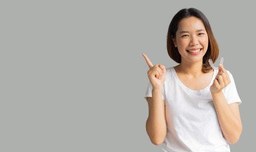 Photo of young Asian woman, smiling, holding a clear aligner while pointing; isolated on gray background