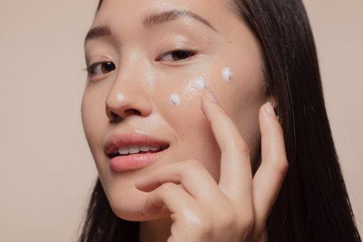 Smiling Asian woman applying dots of facial cream to her face against a beige background.