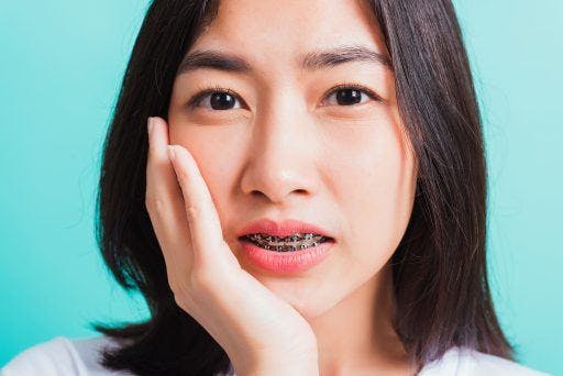Closeup of young Asian girl with braces touching her face against a light blue background. 