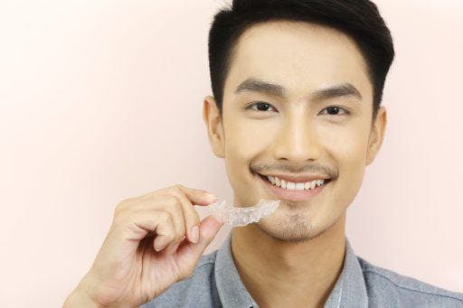 Photo of smiling man holding a clear teeth aligner against a pale pink background