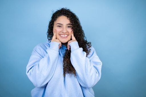 Curly-haired young woman smiling with her gap teeth, fingers on her face. 