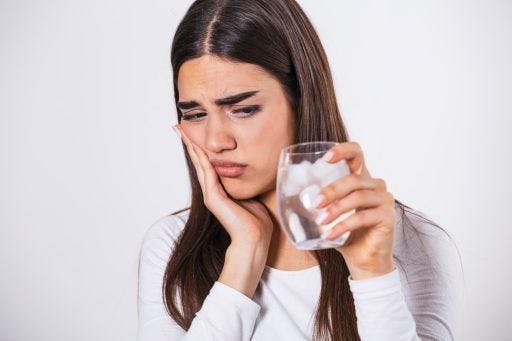 Wincing woman holding the side of her face and a glass of iced water against a white background.