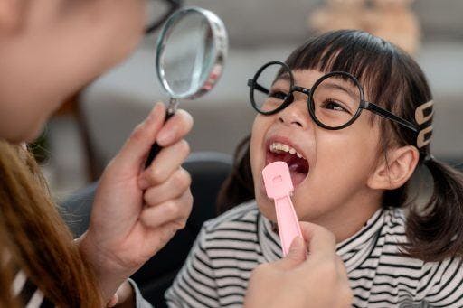 A little girl opens her mouth wide as her mom brushes her tongue and checks her teeth with a magnifying glass.