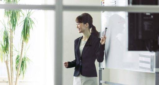 Woman in light grey dress and black blazer giving a presentation with a whiteboard.