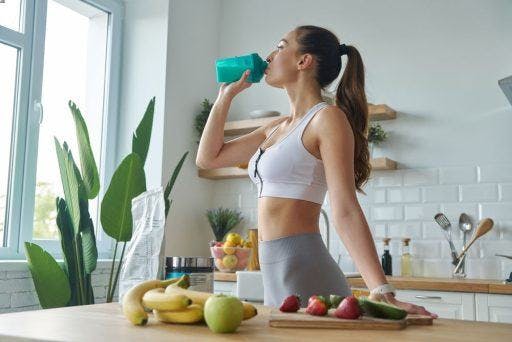 A woman in workout clothes drinking a protein shake and cutting fruits.