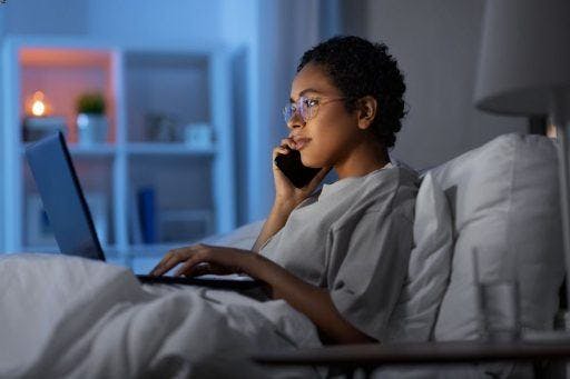 A woman on her phone and laptop in bed, working late at night. 