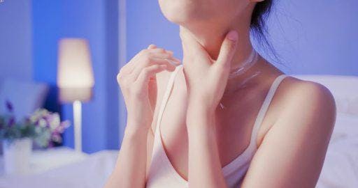 Asian woman in a bedroom applying cream on her neck. 