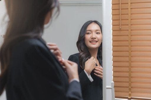 Woman in blazer smiling and looking at herself in the mirror.