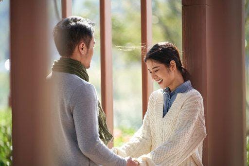 Asian couple in sweaters holding hands by an outdoor gate.