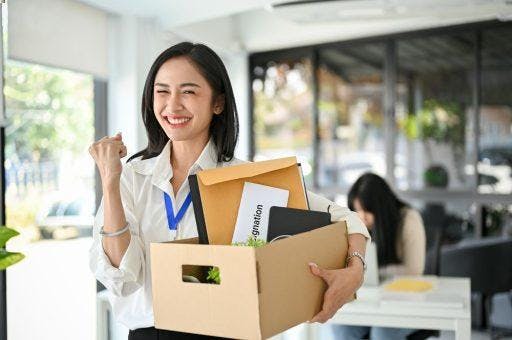 Woman in smiling and putting one hand up while holding a box of office supplies.