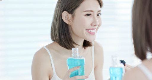 Smiling Asian woman looking in the mirror and holding a bottle of mouthwash in the bathroom.