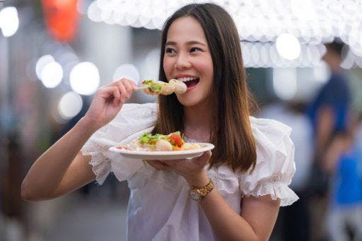 Smiling Asian woman in white eating food on a stick outdoors.
