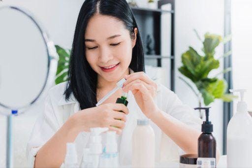 Asian woman in white smiling and applying serum to her hand with visible bottles of skincare in a bright room.