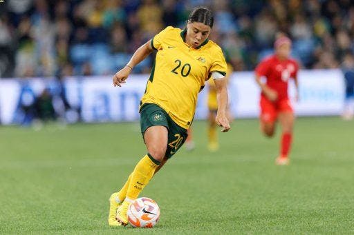 Sam Kerr in a yellow jersey kicking a soccer ball in a field. 