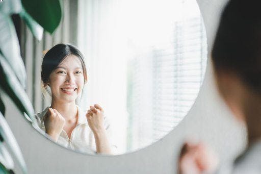 Asian woman smiles confidently and cheers herself on while looking at a mirror.