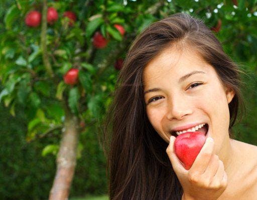 A woman smiling and taking a bite out of an apple outdoors.