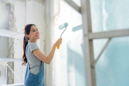 Woman in a grey shirt and overalls painting a wall light blue. 