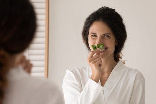 Woman in a white bath robe smiling and smelling a mint leaf in the bathroom.