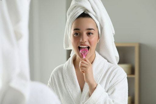 A woman in white robe using a pink tongue scraper in front of the mirror.